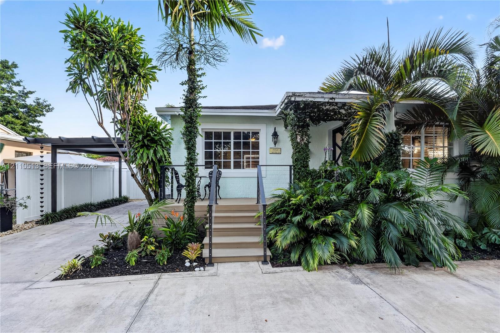 Light-colored exterior siding with grid-pattern windows, raised wood-finish front porch, black metal railing, concrete driveway, and covered carport