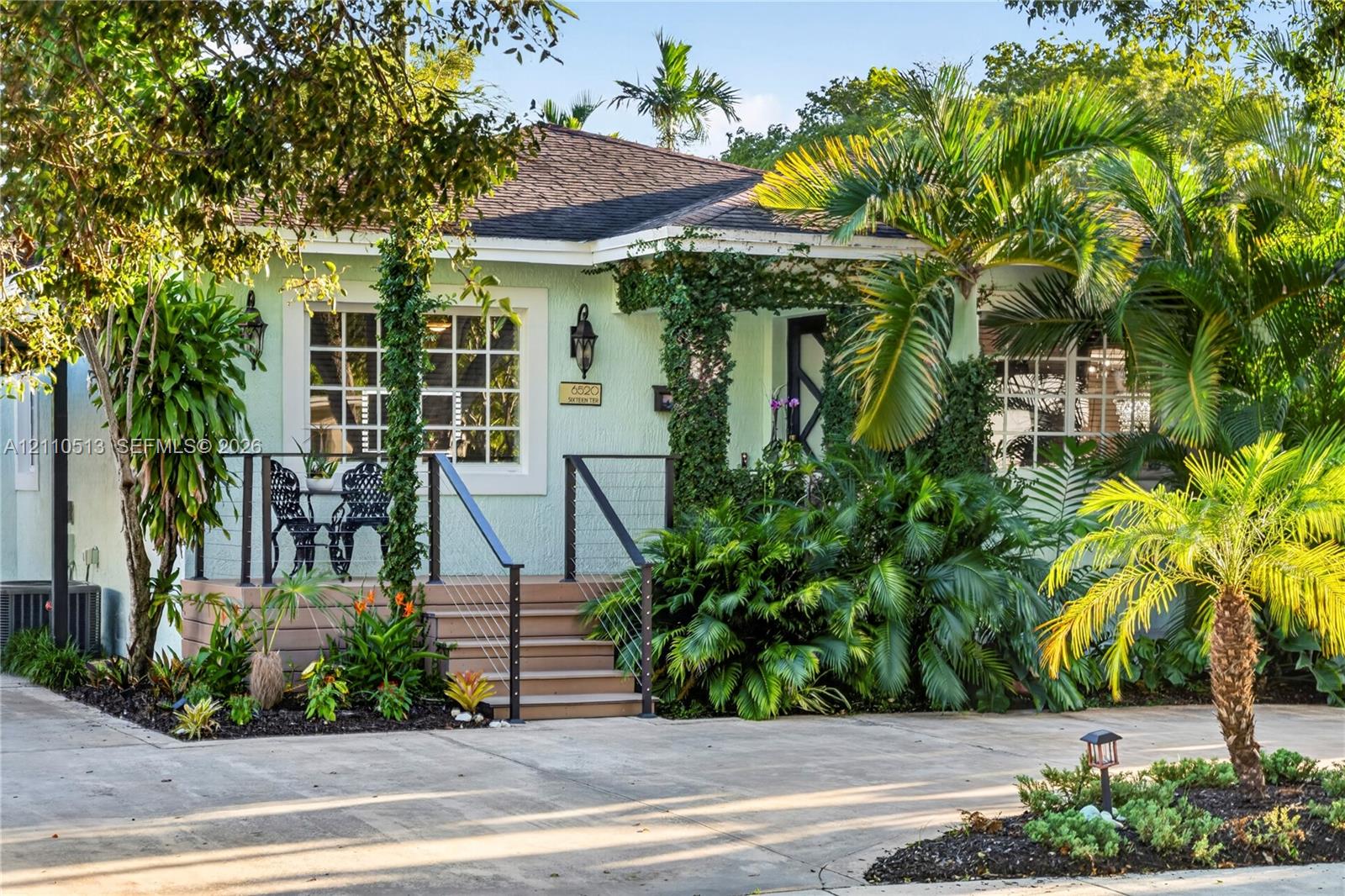 Exterior facade featuring a covered porch with cable railing, shingle roof, multi-pane windows, and extensive mature tropical landscaping