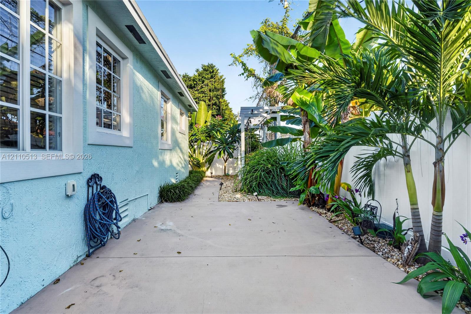 Exterior side yard featuring a concrete walkway, light blue stucco wall, multi-pane windows, privacy fencing, and established tropical landscaping