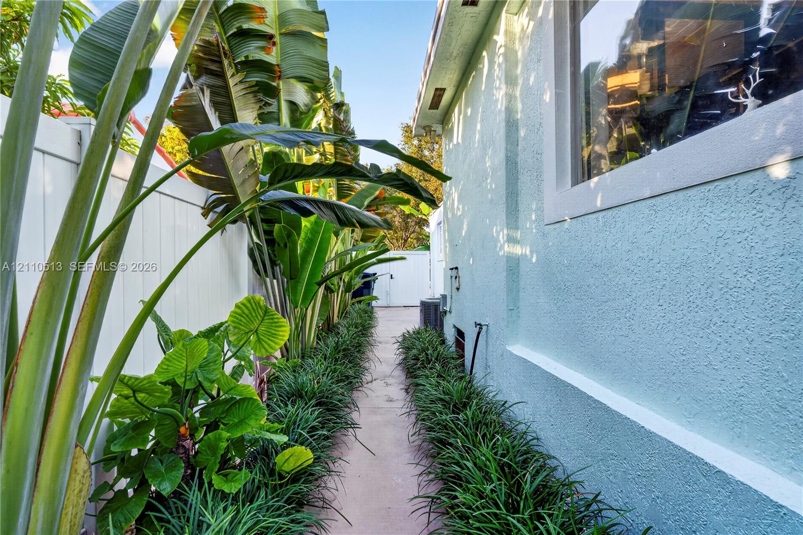 Side yard featuring a concrete pathway, privacy vinyl fencing, mature tropical landscaping, and textured stucco exterior wall