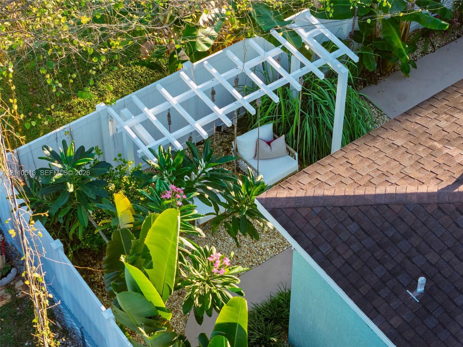 Outdoor area featuring a white vinyl privacy fence, custom pergola, light-toned gravel landscaping, and lush tropical greenery