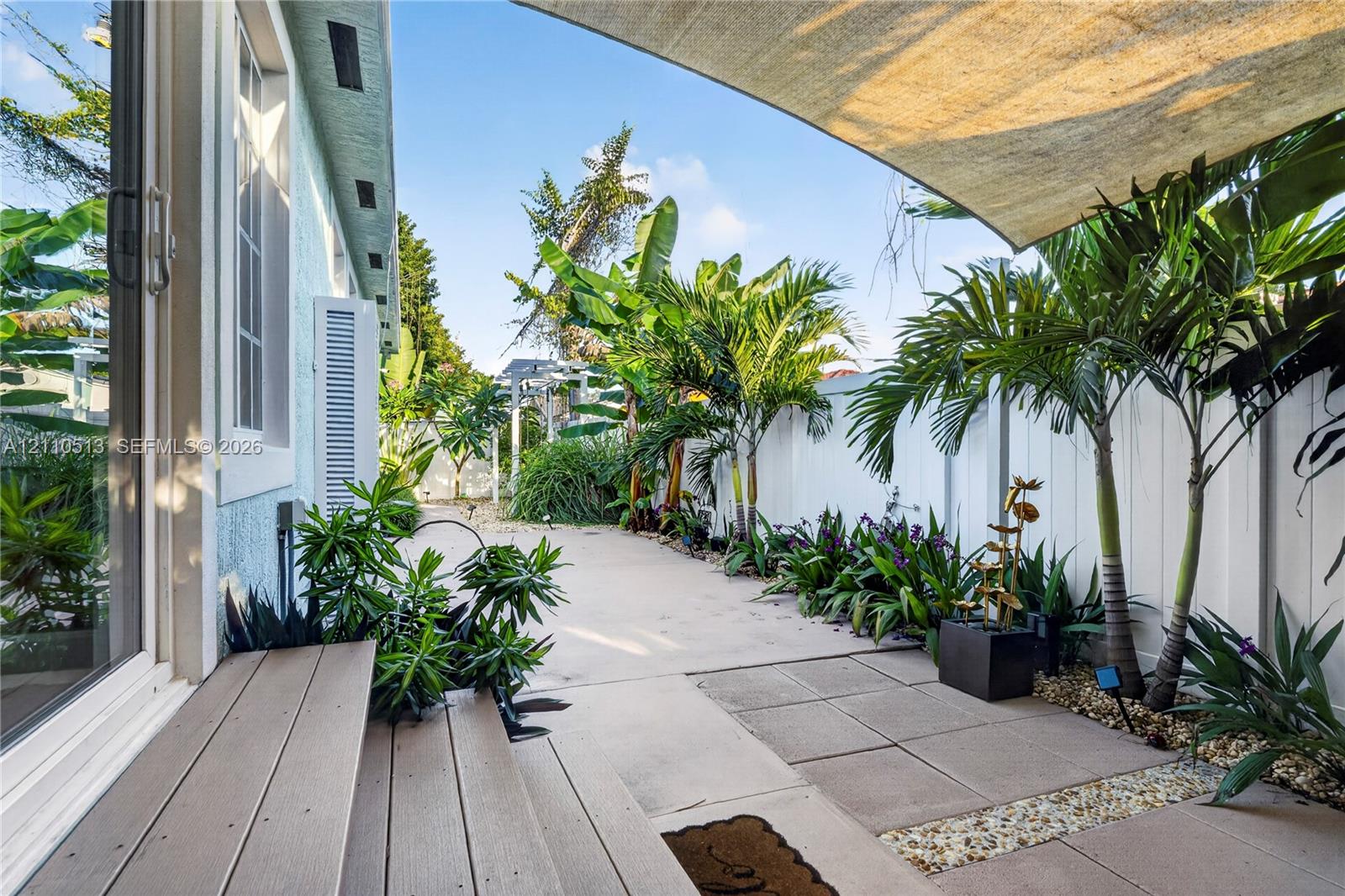 Rear patio featuring a concrete and paved surface, wood-finish decking steps, a retractable shade canopy, white privacy fencing, and mature tropical landscaping