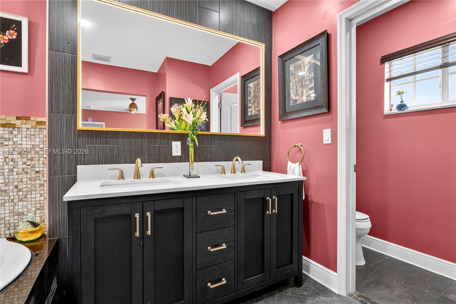 Double vanity featuring a white countertop, dual undermount sinks, gold-tone hardware, and an expansive wall-mounted mirror framed in gold, set against a dark tiled backsplash