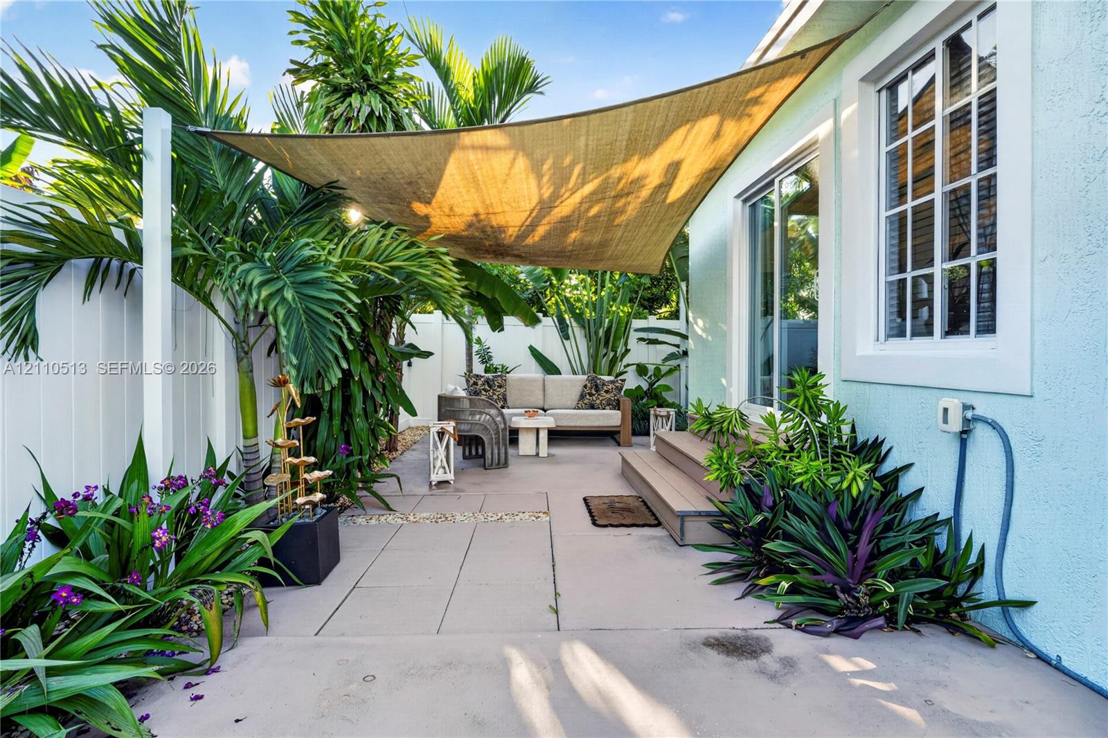 Paved patio area featuring a mounted shade sail, wood-finish stepped platform, light-colored stucco siding, and surrounding privacy fencing