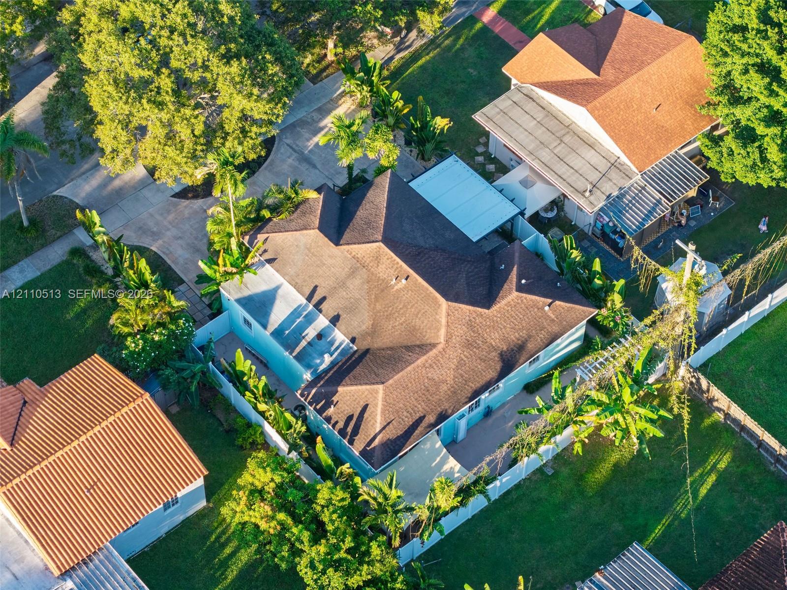 Aerial perspective of a property featuring a multi-pitched shingled roof, a paved driveway, and mature landscaping enclosed by white perimeter fencing Aerial perspective of a property featuring a multi-pitched shingled roof, a paved driveway, and mature landscaping enclosed by white perimeter fencing