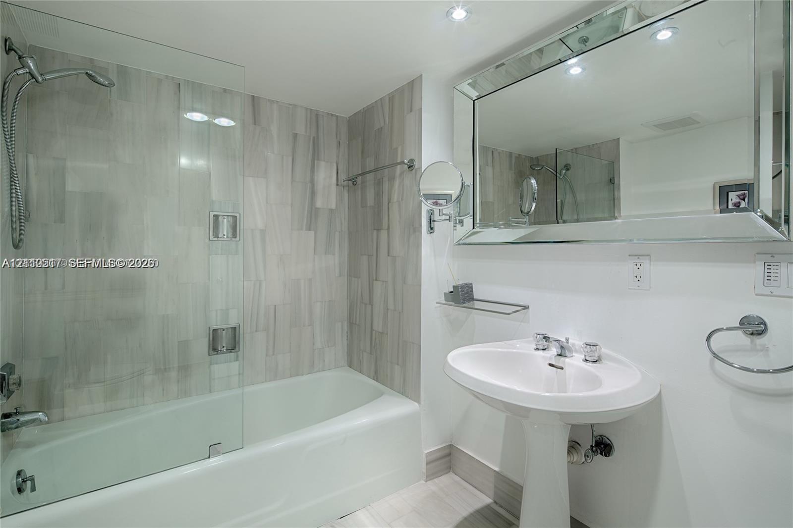 Bathroom featuring a white pedestal sink, frameless glass shower screen, tub with wall-mounted shower head, large beveled mirror, and stone-finish wall tile Bathroom featuring a white pedestal sink, frameless glass shower screen, tub with wall-mounted shower head, large beveled mirror, and stone-finish wall tile