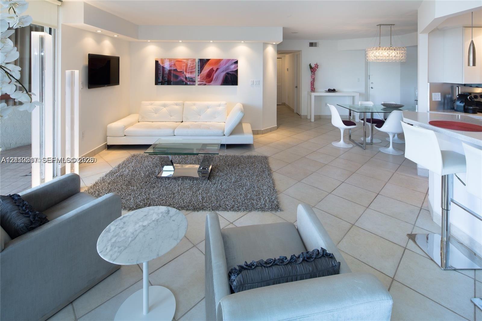 Open concept living area featuring large-format ceramic tile flooring, recessed ceiling lighting, a breakfast bar with stone-finish countertop, and a tiered crystal chandelier Open concept living area featuring large-format ceramic tile flooring, recessed ceiling lighting, a breakfast bar with stone-finish countertop, and a tiered crystal chandelier