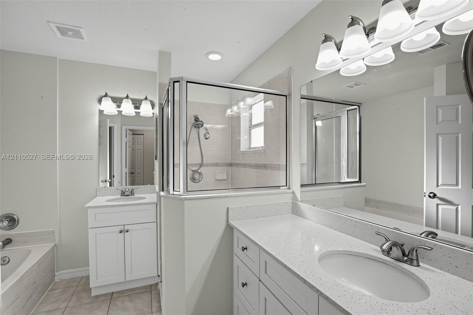Bathroom featuring dual white shaker-style cabinetry, light-toned stone-finish countertops, an enclosed glass-paneled shower with tiled surround, and large-scale vanity mirrors with multi-bulb light fixtures