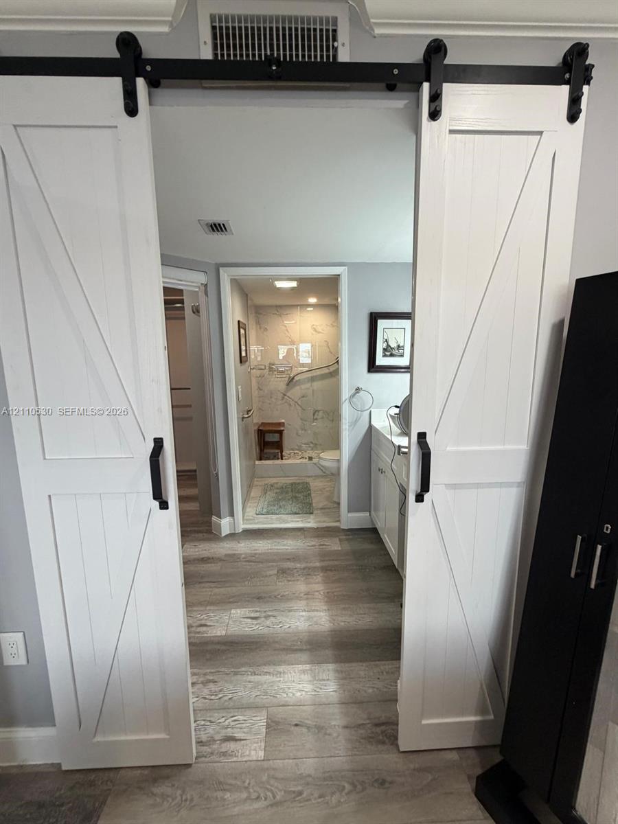 White barn-style sliding doors with black hardware, wood-finish flooring, white cabinetry, and shower enclosure with wall-mounted grab bars