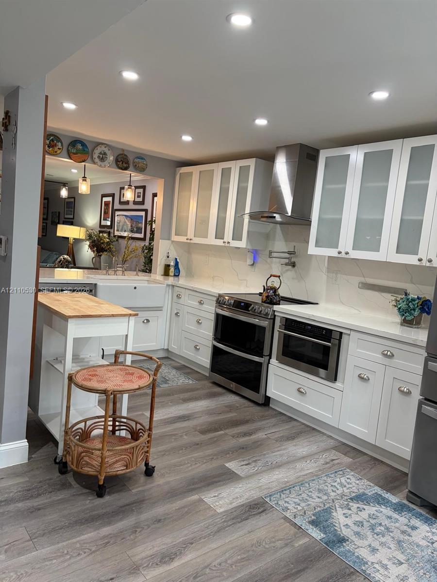 Kitchen featuring white cabinetry with glass-front uppers, stone-finish countertops, apron-front sink, stainless steel range hood, and wood-finish flooring