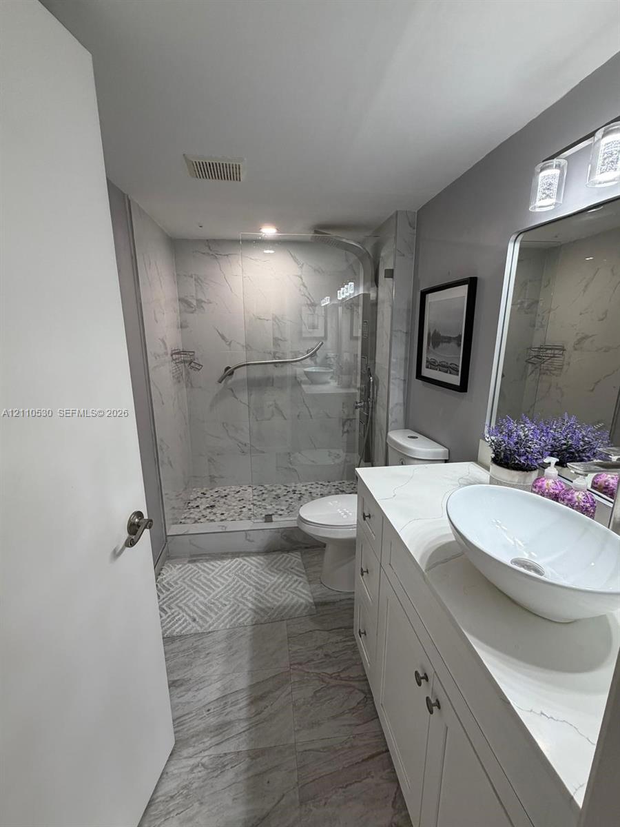 Bathroom featuring a white vanity with a rectangular vessel sink and solid-surface countertop, walk-in shower with stone-patterned wall tile and pebble-texture flooring, and large-format tile flooring