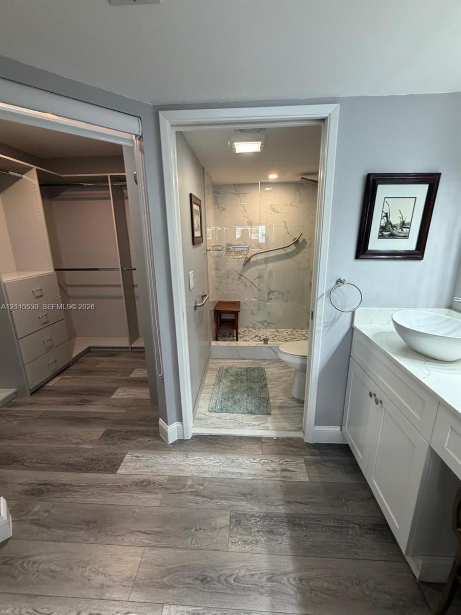 Bathroom featuring a white vanity with a vessel sink, wood-finish flooring, and a walk-in shower with stone-look wall tile and pebble-tile flooring, paired with a custom-built walk-in closet