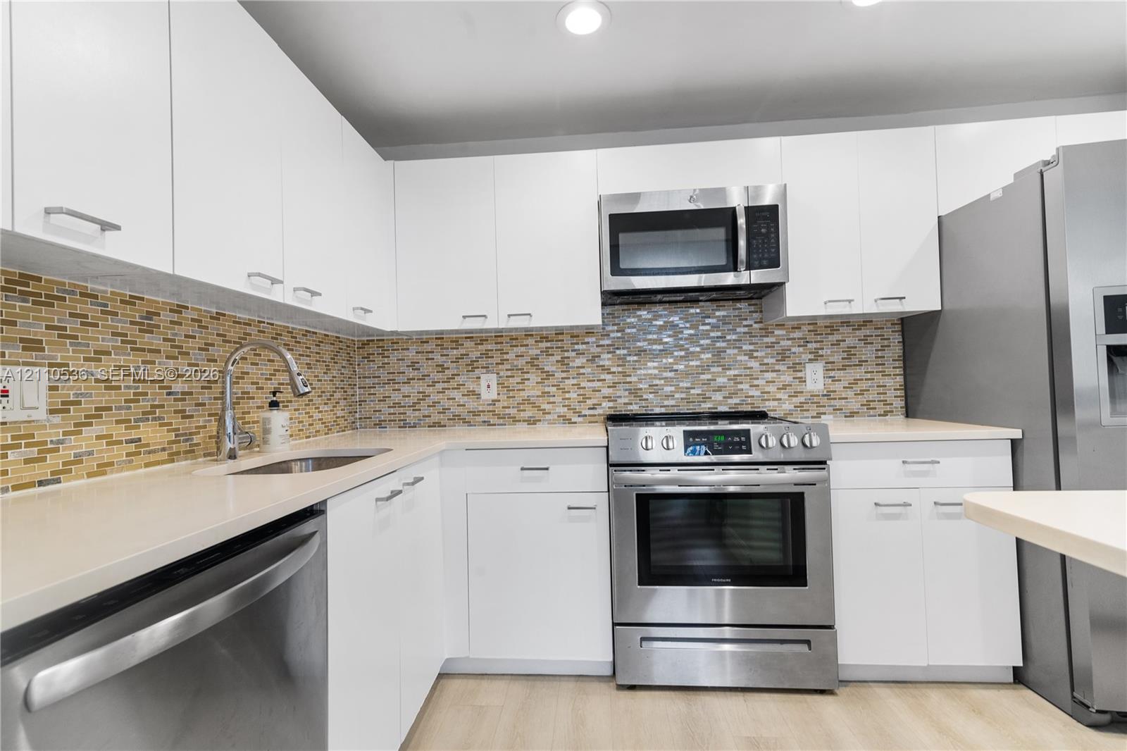 Kitchen featuring flat-panel white cabinetry, stainless steel appliances, multi-tonal mosaic tile backsplash, solid-surface countertops, and wood-finish flooring Kitchen featuring flat-panel white cabinetry, stainless steel appliances, multi-tonal mosaic tile backsplash, solid-surface countertops, and wood-finish flooring