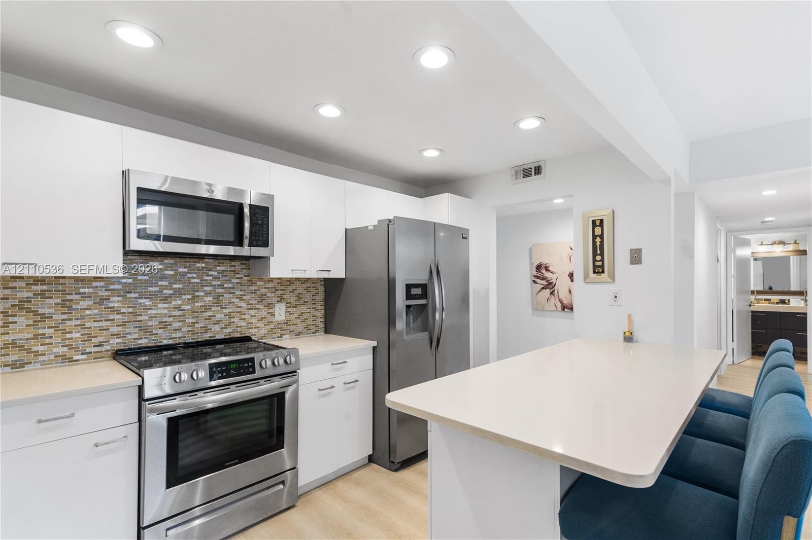 Kitchen featuring flat-panel white cabinetry, stainless steel appliances, multi-tonal glass tile backsplash, light-toned countertop, and recessed lighting Kitchen featuring flat-panel white cabinetry, stainless steel appliances, multi-tonal glass tile backsplash, light-toned countertop, and recessed lighting