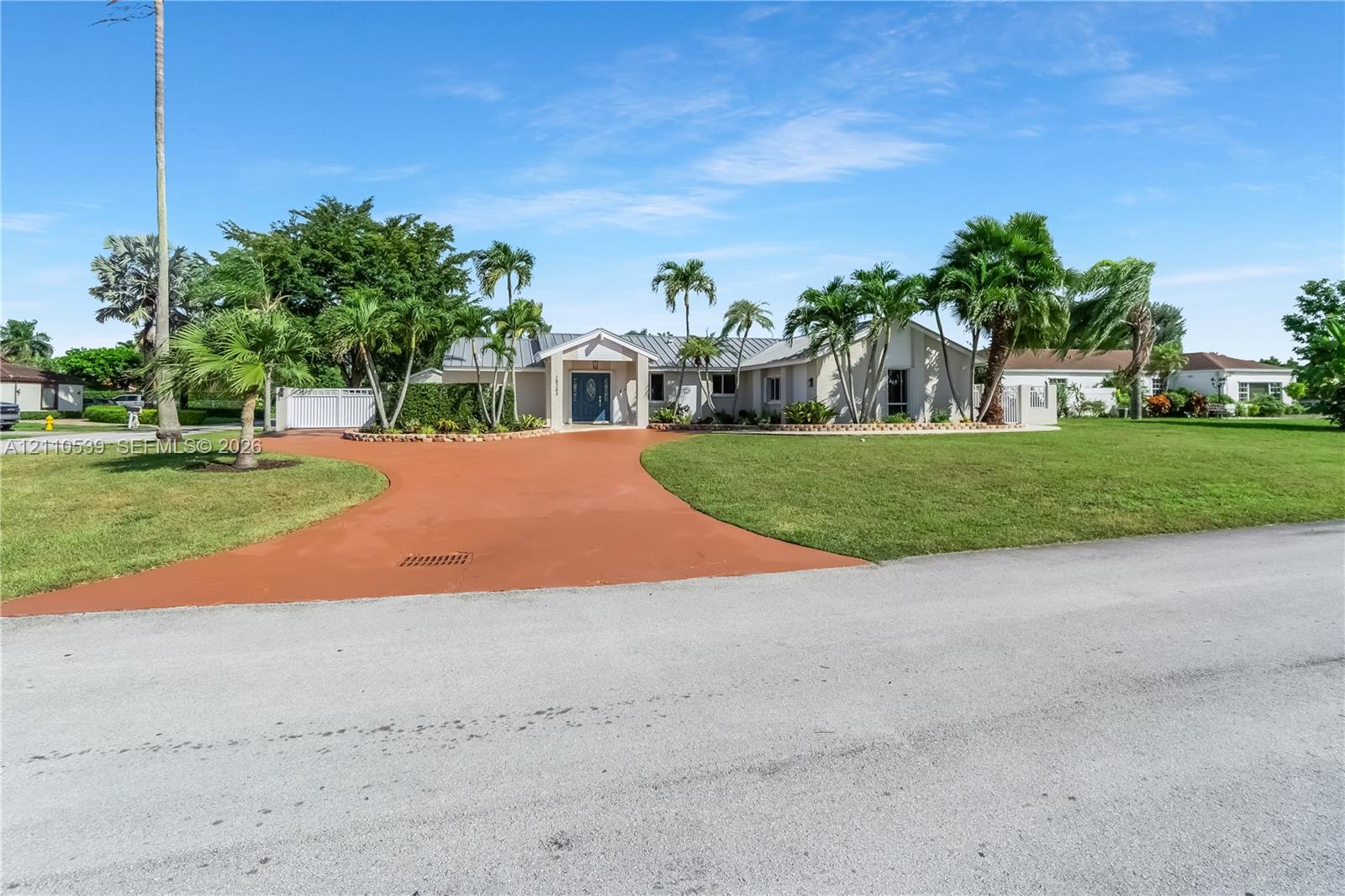 Single-story residence featuring a metal roof, an expansive stained concrete driveway, mature palm trees, and a gated side perimeter