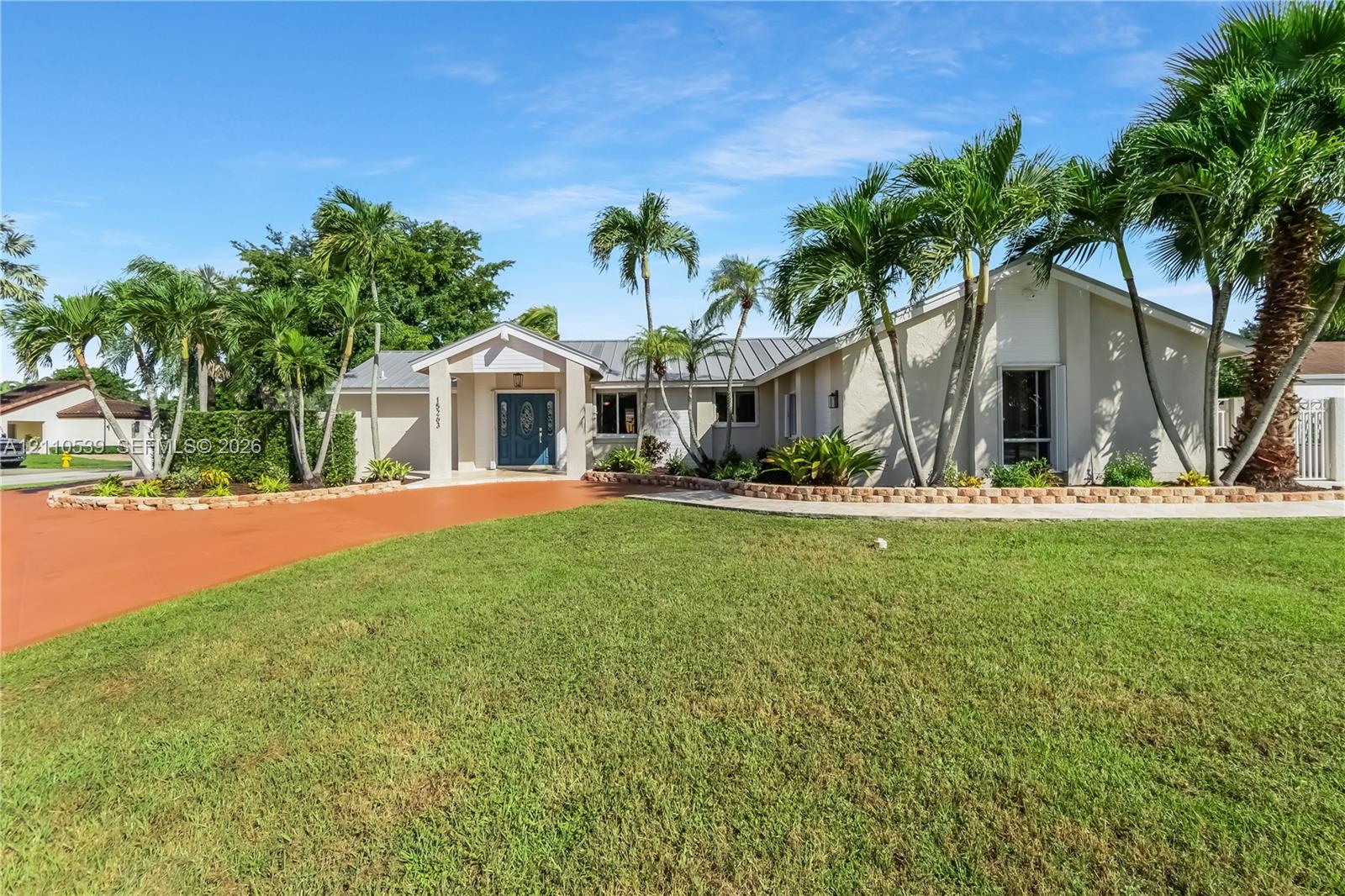 Single-story residence featuring a painted stucco exterior, metal roofing, expansive front lawn, terracotta-toned driveway, and manicured palm landscaping