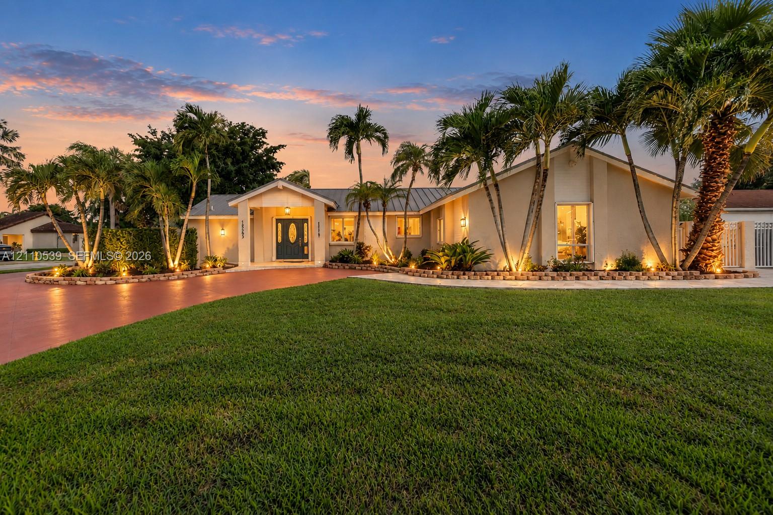 Stucco exterior featuring a covered entryway, metal roofing, brick-edged landscaping, mature palm trees, and a paved driveway