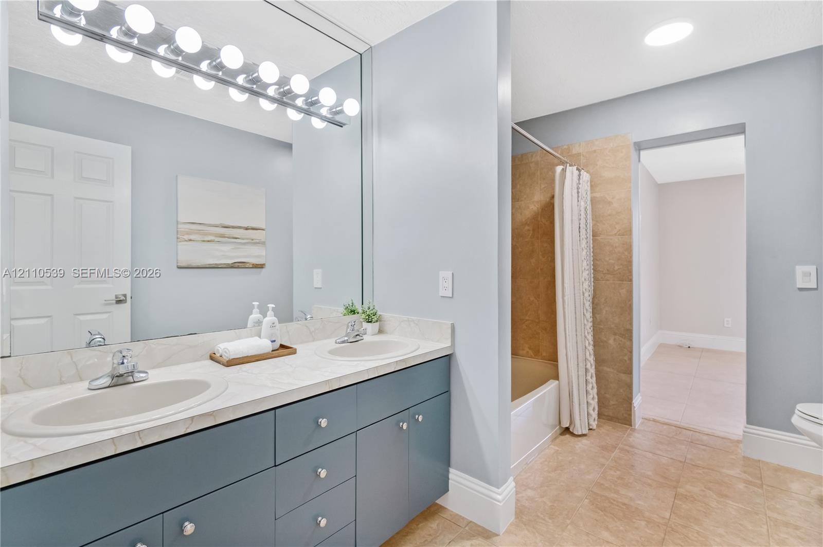 Dual-sink vanity with a solid-surface countertop and recessed basins, expansive wall-mounted vanity mirror, multi-bulb overhead lighting, and tub-shower combination featuring neutral-toned tile surround