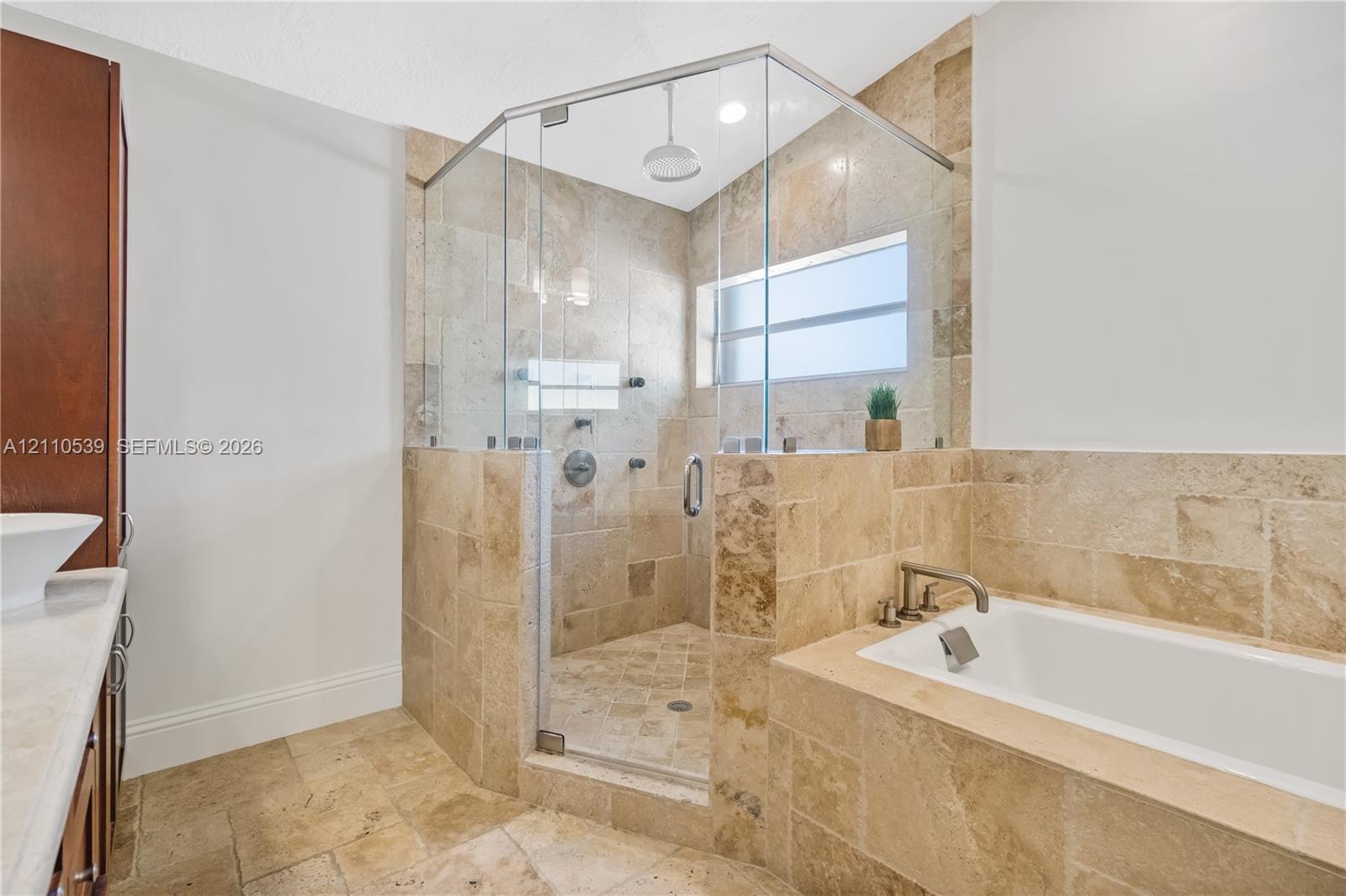 Walk-in shower with a glass enclosure, stone-tile surround, and ceiling-mounted rainfall showerhead, accompanied by a built-in soaking tub with a stone-tile deck and polished metal fixtures