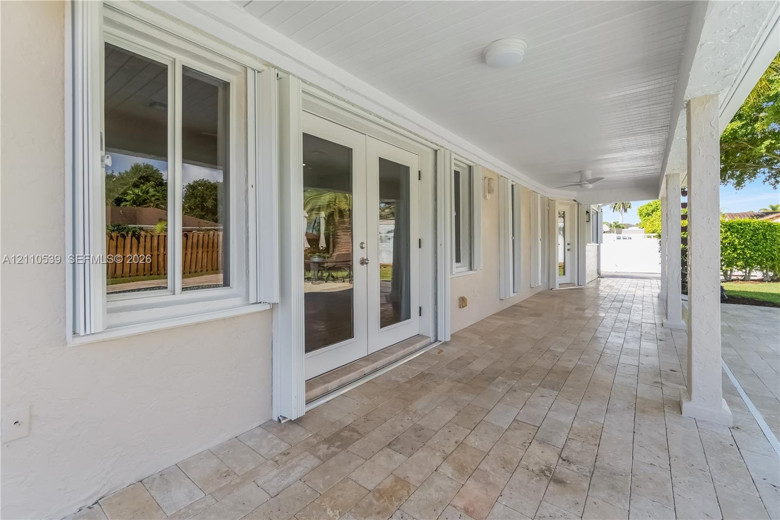 Covered patio featuring stone-tile flooring, glass-paned double doors, white-trimmed windows, and ceiling-mounted light fixtures