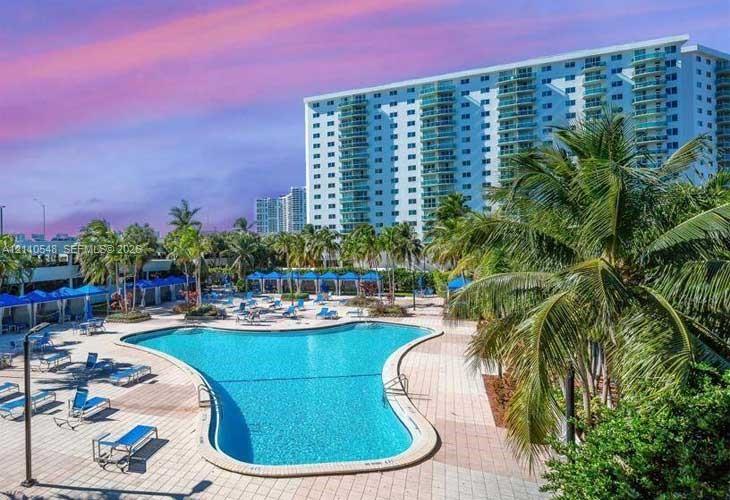 Outdoor swimming pool surrounded by a paved deck, shaded cabanas, and mature palm trees