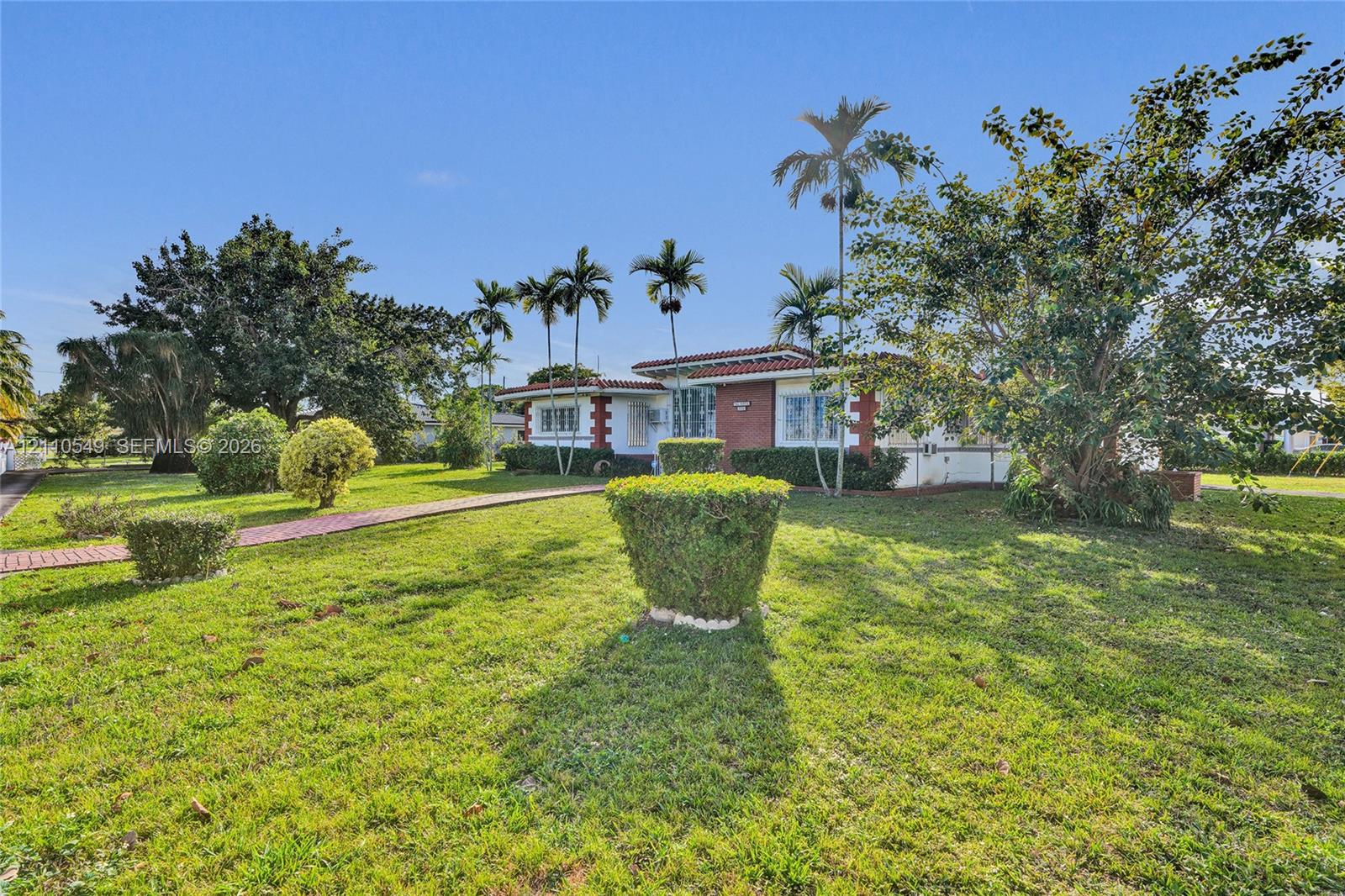 Single-story residence featuring a tile roof, brick exterior accents, multi-pane windows, and a paved walkway situated on a lot with mature palm trees