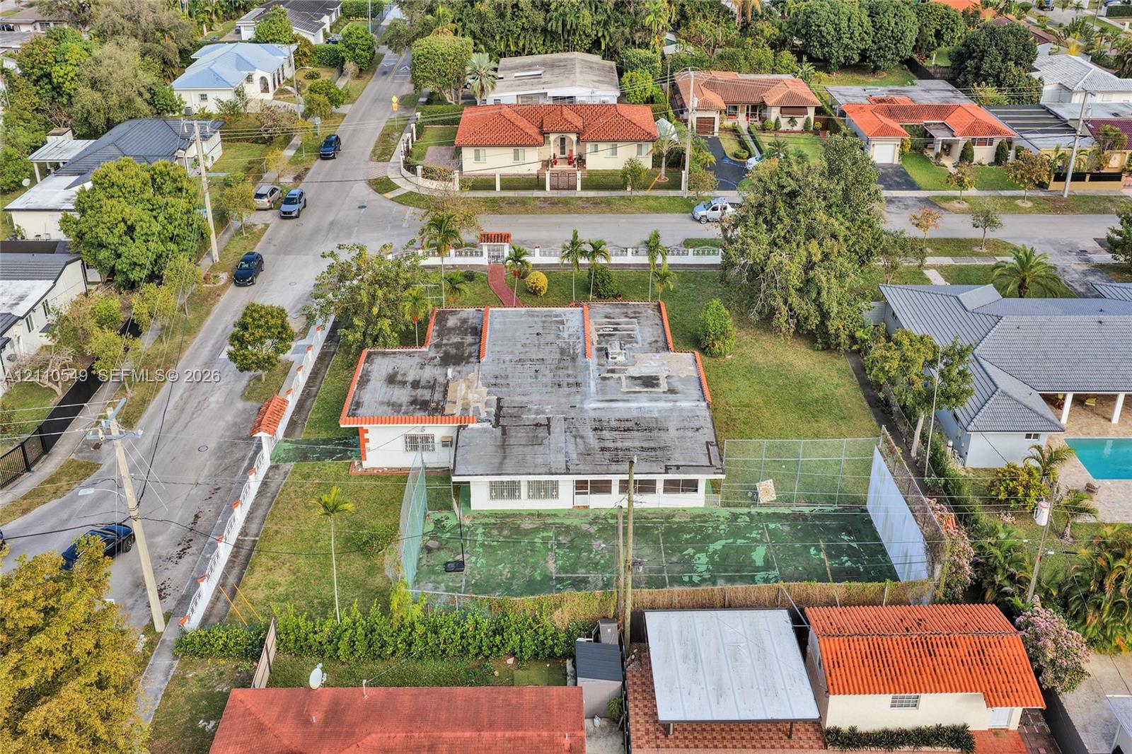 Aerial perspective of a corner lot property featuring a single-story residence, a private fenced-in sport court, expansive green space, and mature landscaping