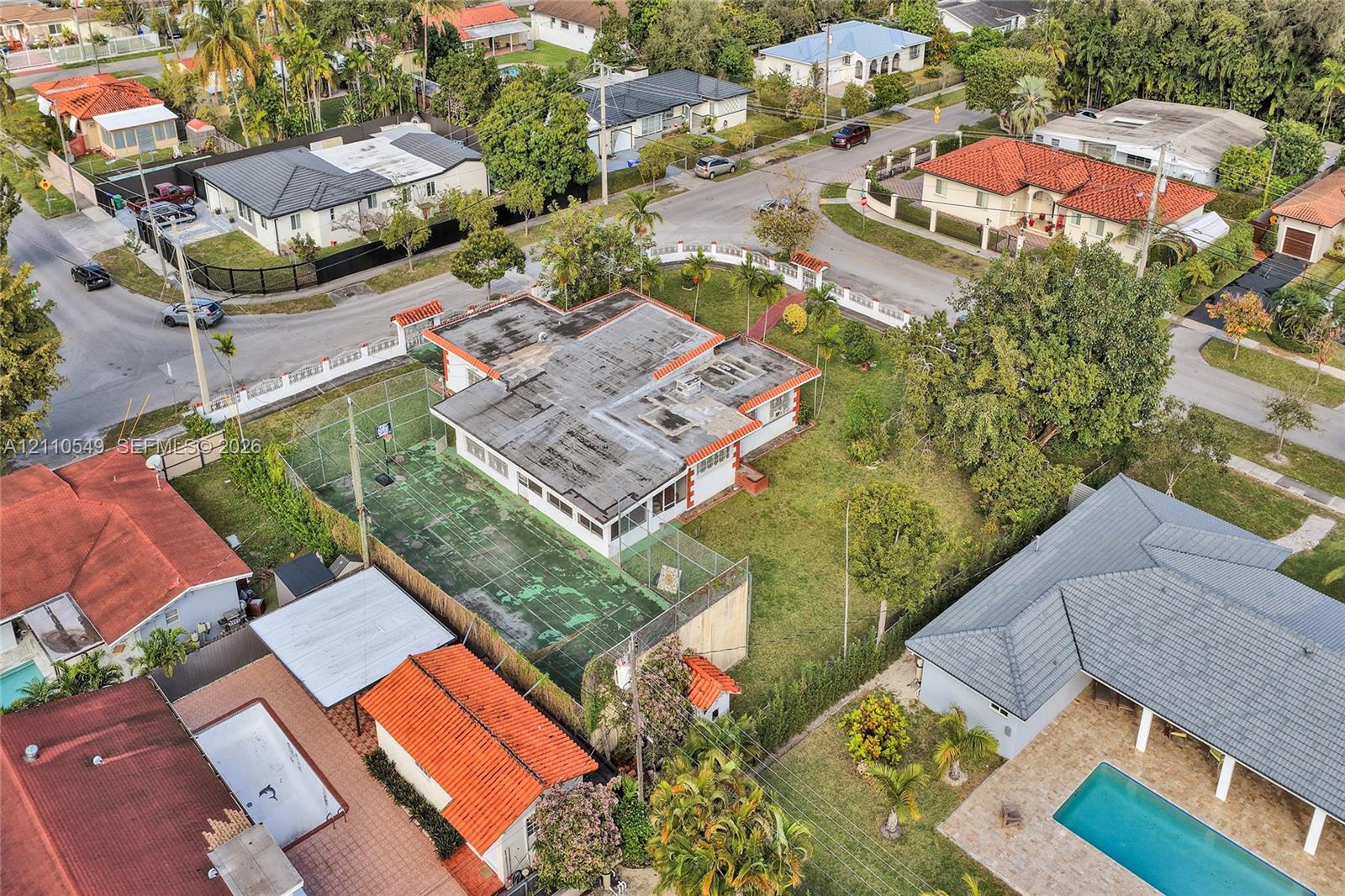 Aerial perspective of a corner lot property featuring a private fenced sports court, expansive green space, and mature perimeter trees