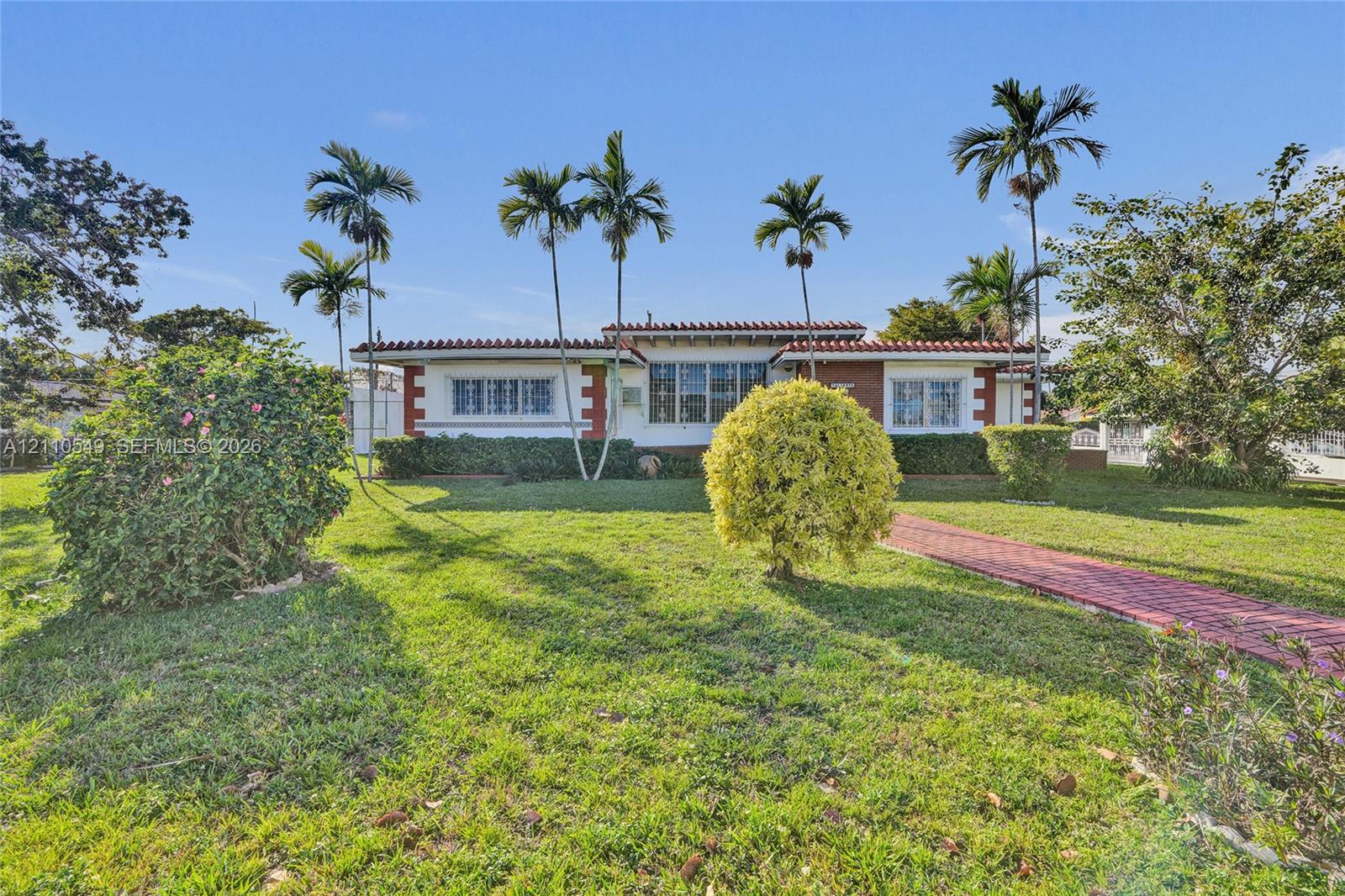 Single-story exterior featuring a clay tile roof, brick accents, multi-pane windows, and a paved brick walkway set within a landscaped lawn with mature palm trees