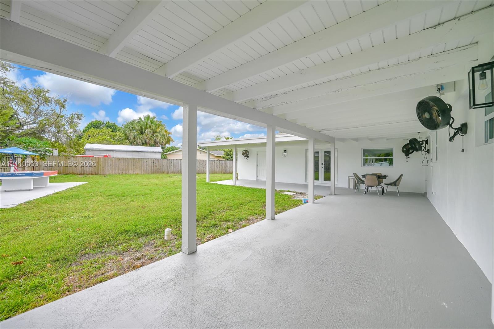 Covered rear patio featuring a painted concrete floor, wood-beam ceiling, wall-mounted outdoor lighting, and expansive backyard space enclosed by wood fencing