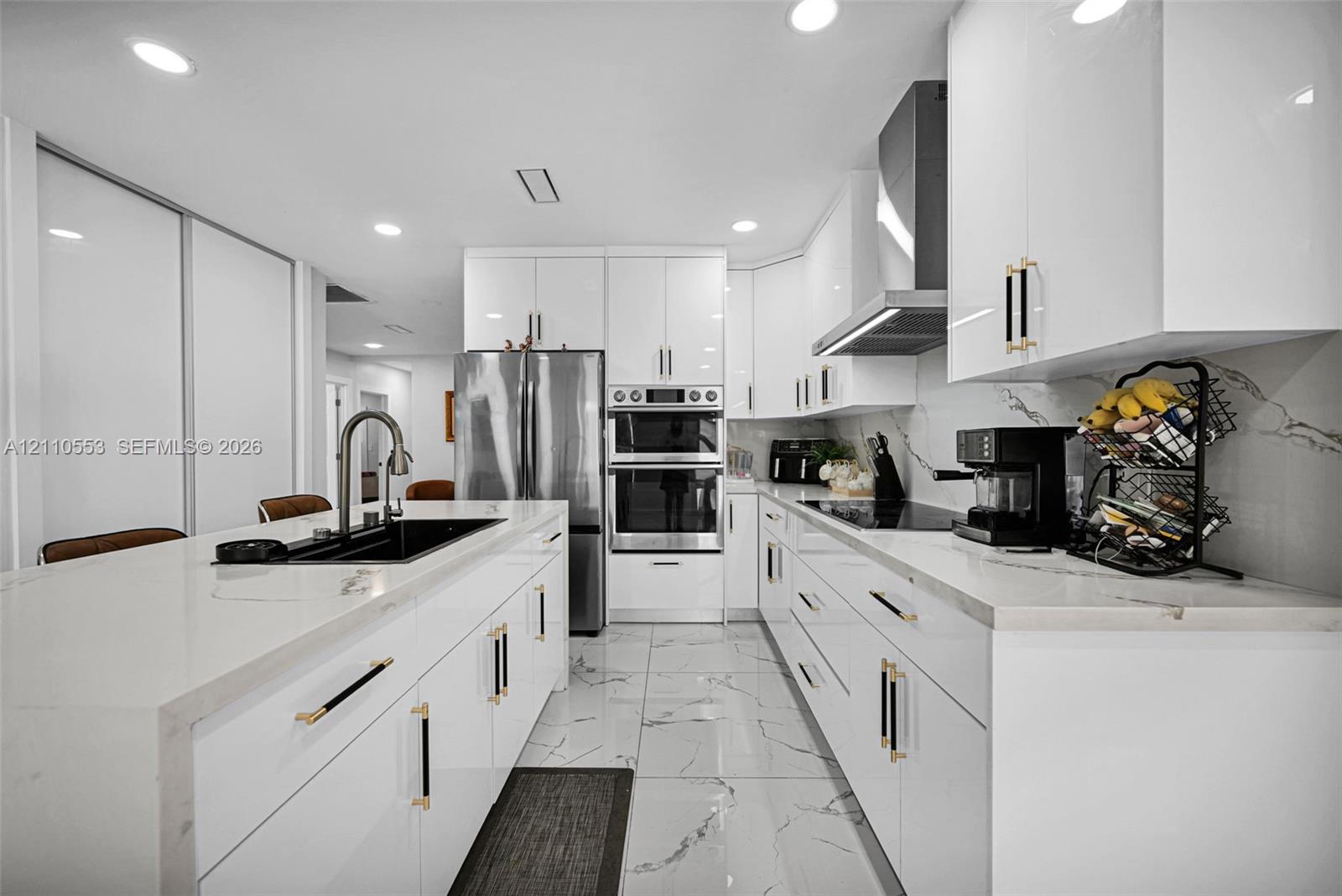 Modern kitchen featuring high-gloss white cabinetry, white stone-finish countertops, large-format veined tile flooring, built-in double wall ovens, and a center island with a matte black undermount sink