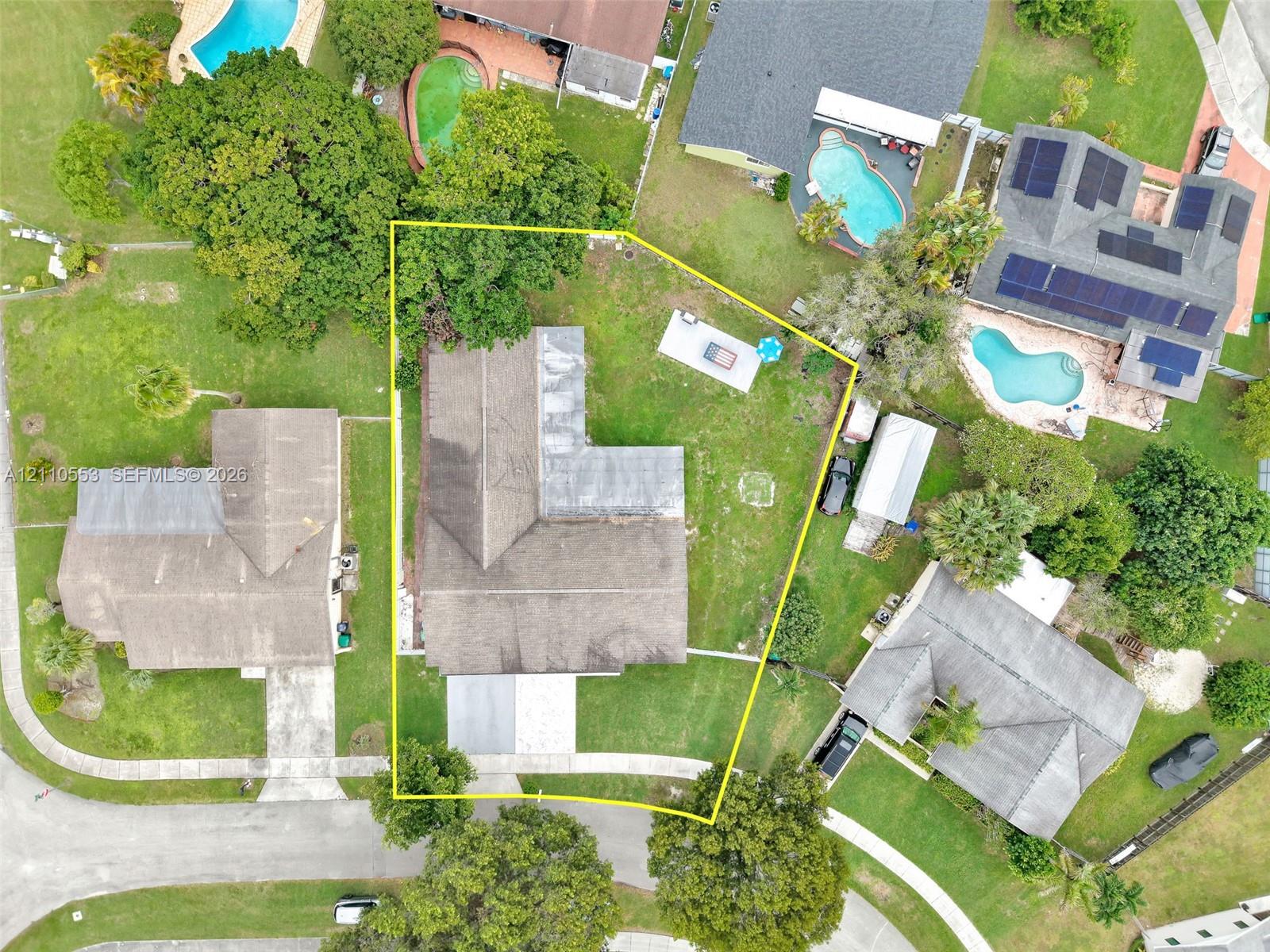Aerial perspective of the property featuring a structured lot with an L-shaped roofline, mature trees, and a paved driveway