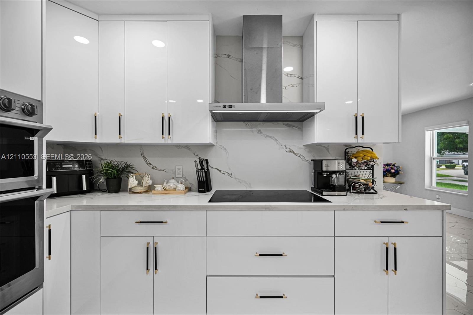 Kitchen featuring glossy white flat-panel cabinetry, white stone-patterned backsplash, stainless steel chimney-style range hood, electric cooktop, and built-in double wall oven