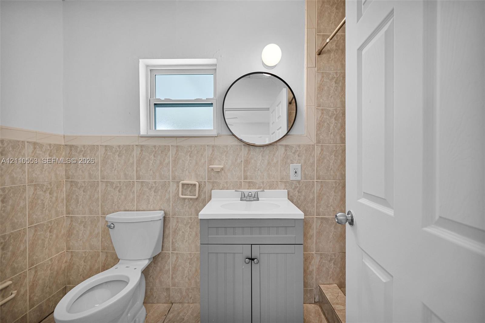 Bathroom featuring neutral-toned wall tile, a gray vanity cabinet with a white countertop, a circular wall-mounted mirror, and a single-hung window
