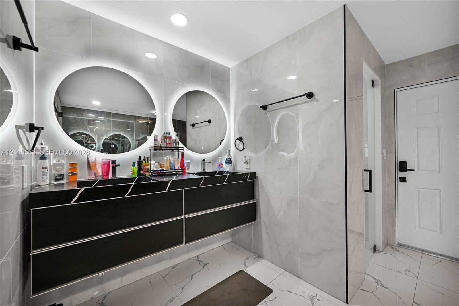 Floating dual-sink vanity with matte black finish, integrated backlit circular mirrors, large-format polished wall tile, and light-toned floor tile with dark veining