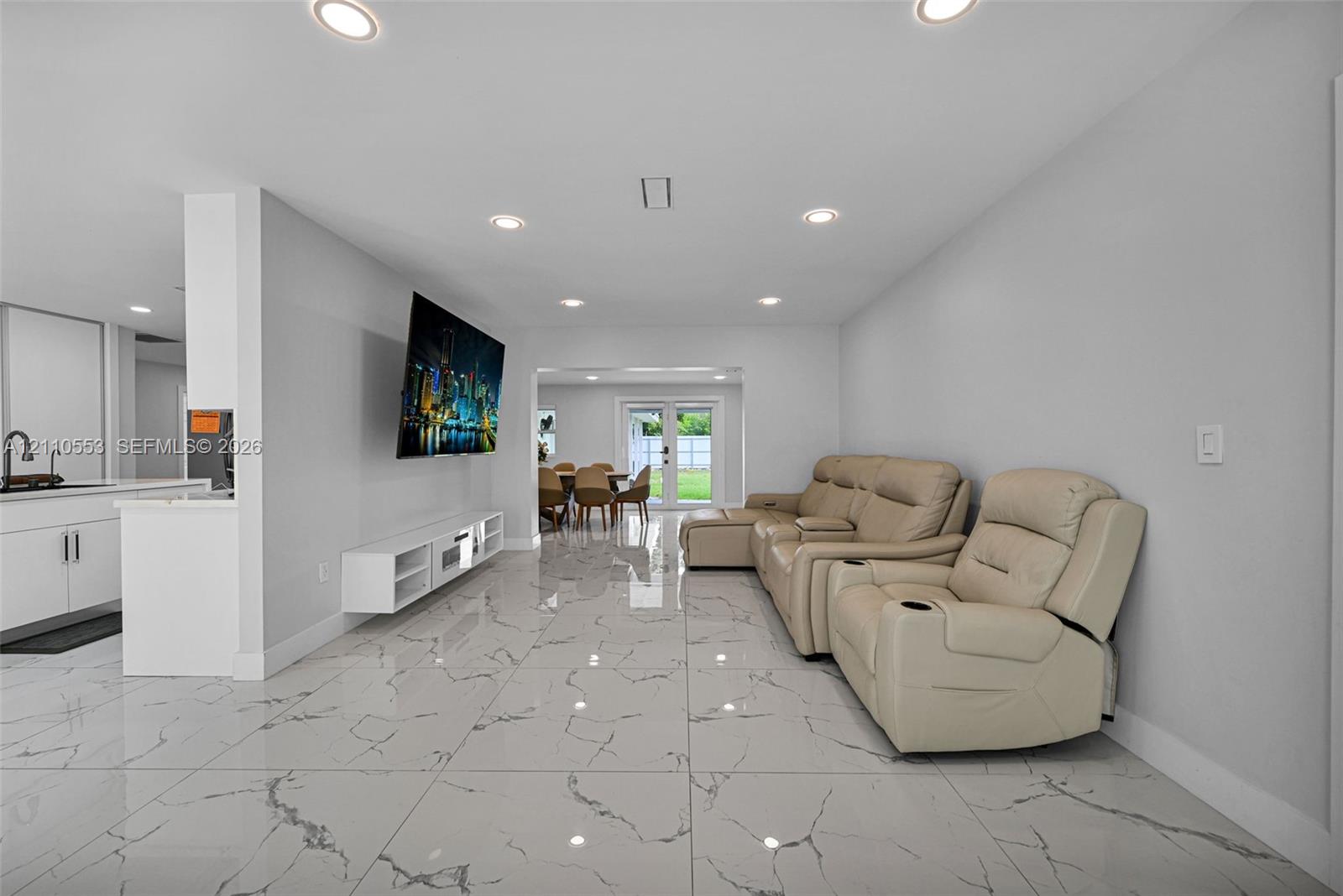 Open-concept interior featuring large-format stone-patterned tile flooring, recessed ceiling lighting, a wall-mounted media console, and dual-pane glass entry doors