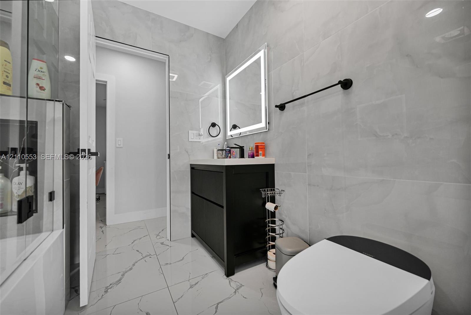 Bathroom featuring large-format porcelain tile wall cladding, a black floating vanity with a solid surface countertop, an LED-backlit mirror, and matte black hardware