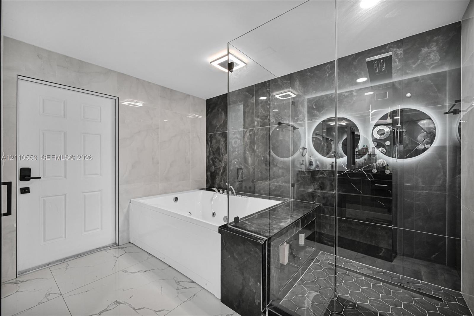 Jetted soaking tub, walk-in shower with glass enclosure, large-format tile flooring, polished stone wall surfacing, and dual illuminated circular mirrors