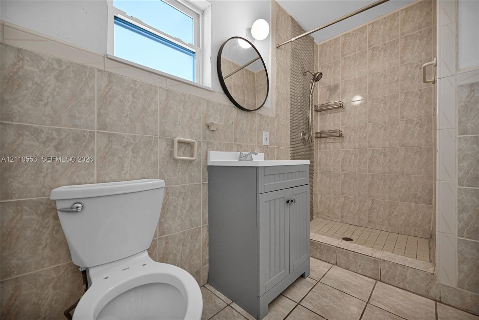Bathroom featuring tile-surround walk-in shower, grey shaker-style vanity with white countertop, round wall-mounted mirror, and tile flooring