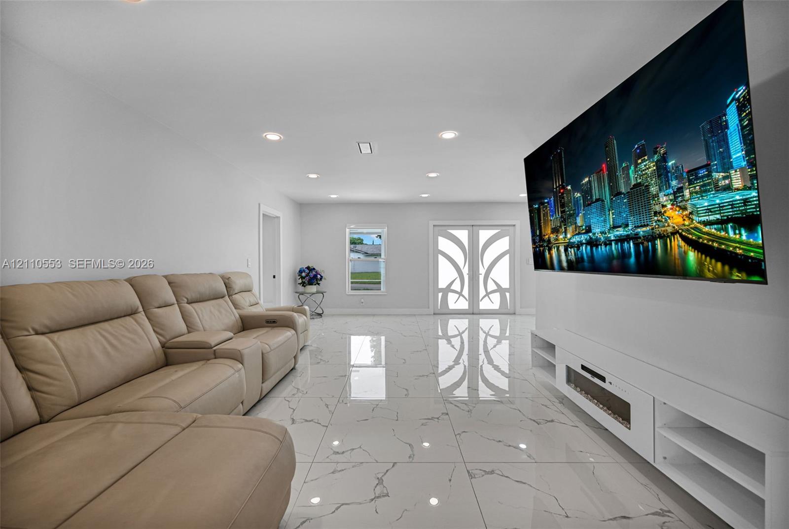 Large-format tile flooring with marble-style veining, recessed lighting, double entry doors with decorative glass inserts, and a white built-in media console with an integrated electric fireplace