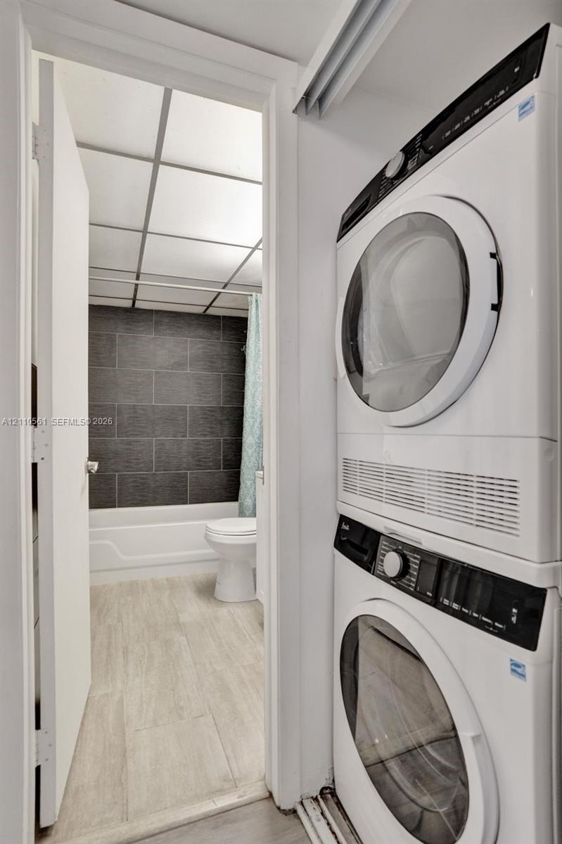 Stacked laundry unit featuring a front-loading washer and dryer, situated near a bathroom with wood-finish flooring, a white bathtub, and dark horizontal wall tiling