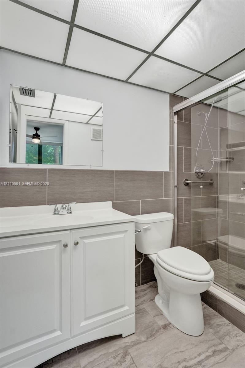 Bathroom featuring a white vanity cabinet, solid-surface countertop, walk-in shower with tiled walls, and drop ceiling panel lighting