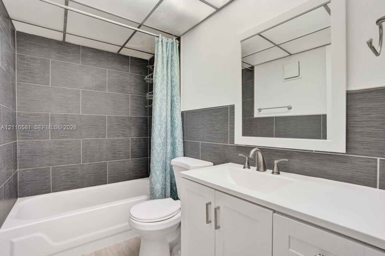 Bathroom featuring a white vanity cabinet with a solid surface countertop, an undermount sink, brushed nickel hardware, and grey textured tile surround in the shower area