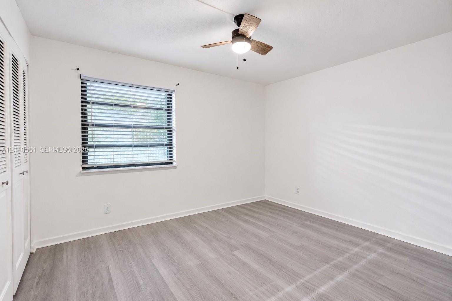 Neutral-toned wood-finish flooring, ceiling fan with integrated lighting, window with horizontal blinds, and double-door closet with louvered panels