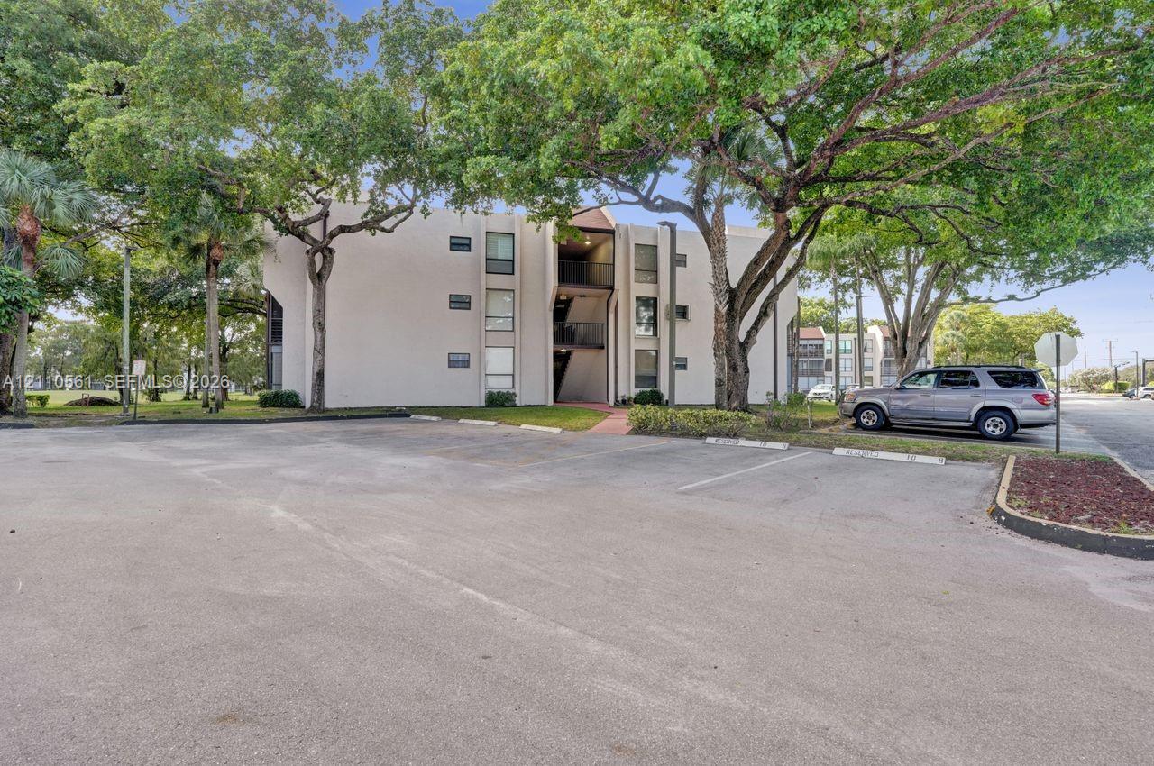 Multi-story building with light-toned facade, asphalt parking area, and mature canopy trees