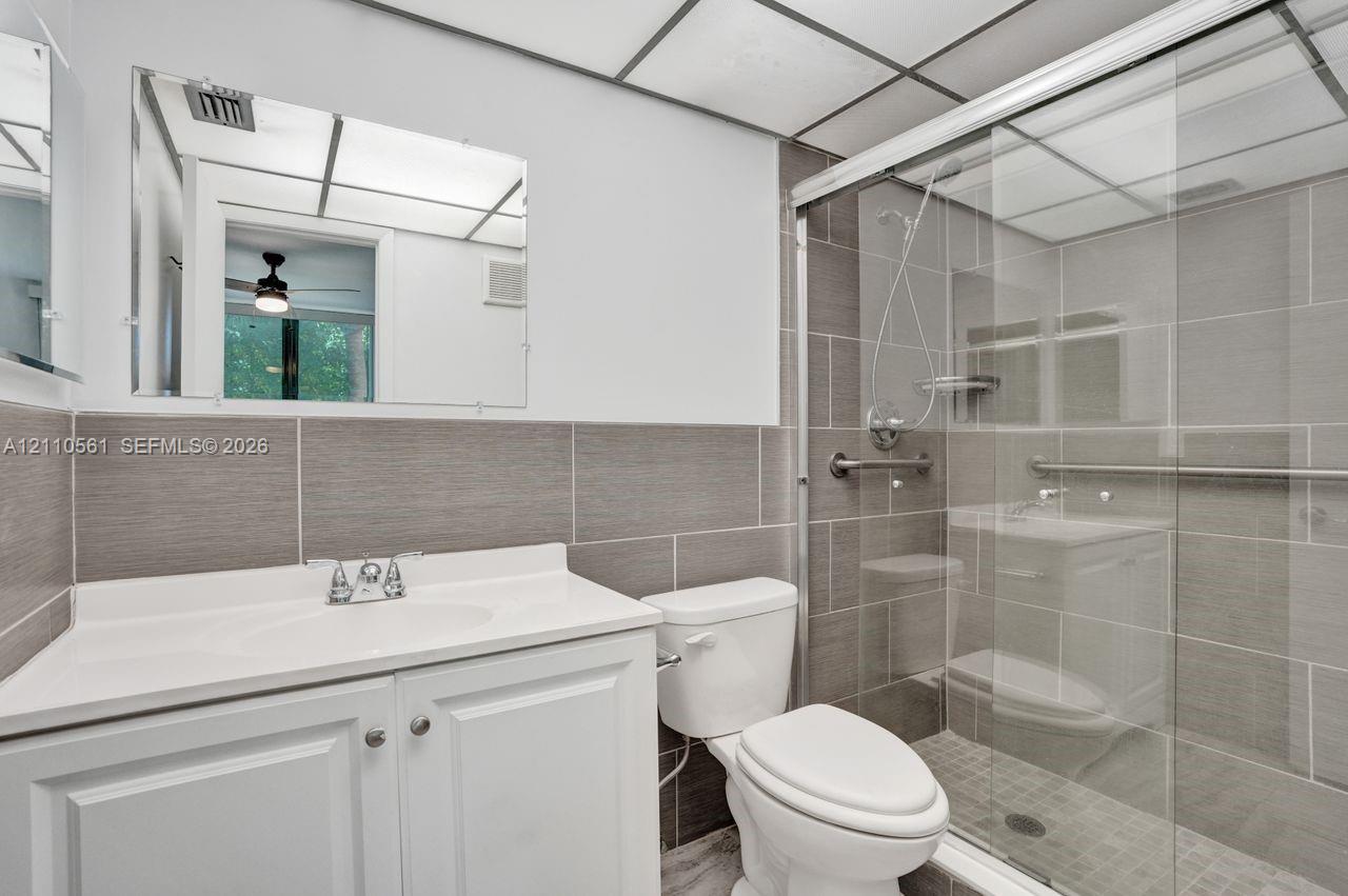 Bathroom featuring a white vanity cabinet with an integrated sink, rectangular tile surround, glass-enclosed shower, and grid-patterned ceiling panels