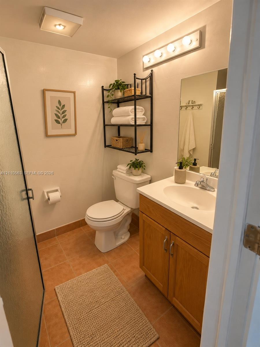 Bathroom featuring wood-finish vanity cabinetry with an integrated sink, tiled flooring, wall-mounted light fixture, and walk-in shower with a glass door Bathroom featuring wood-finish vanity cabinetry with an integrated sink, tiled flooring, wall-mounted light fixture, and walk-in shower with a glass door
