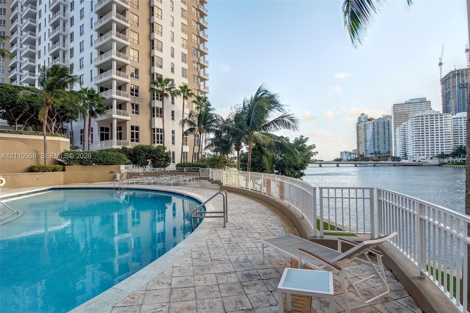 Curved swimming pool with stone-tiled deck, white metal perimeter fencing, and palms overlooking the waterway