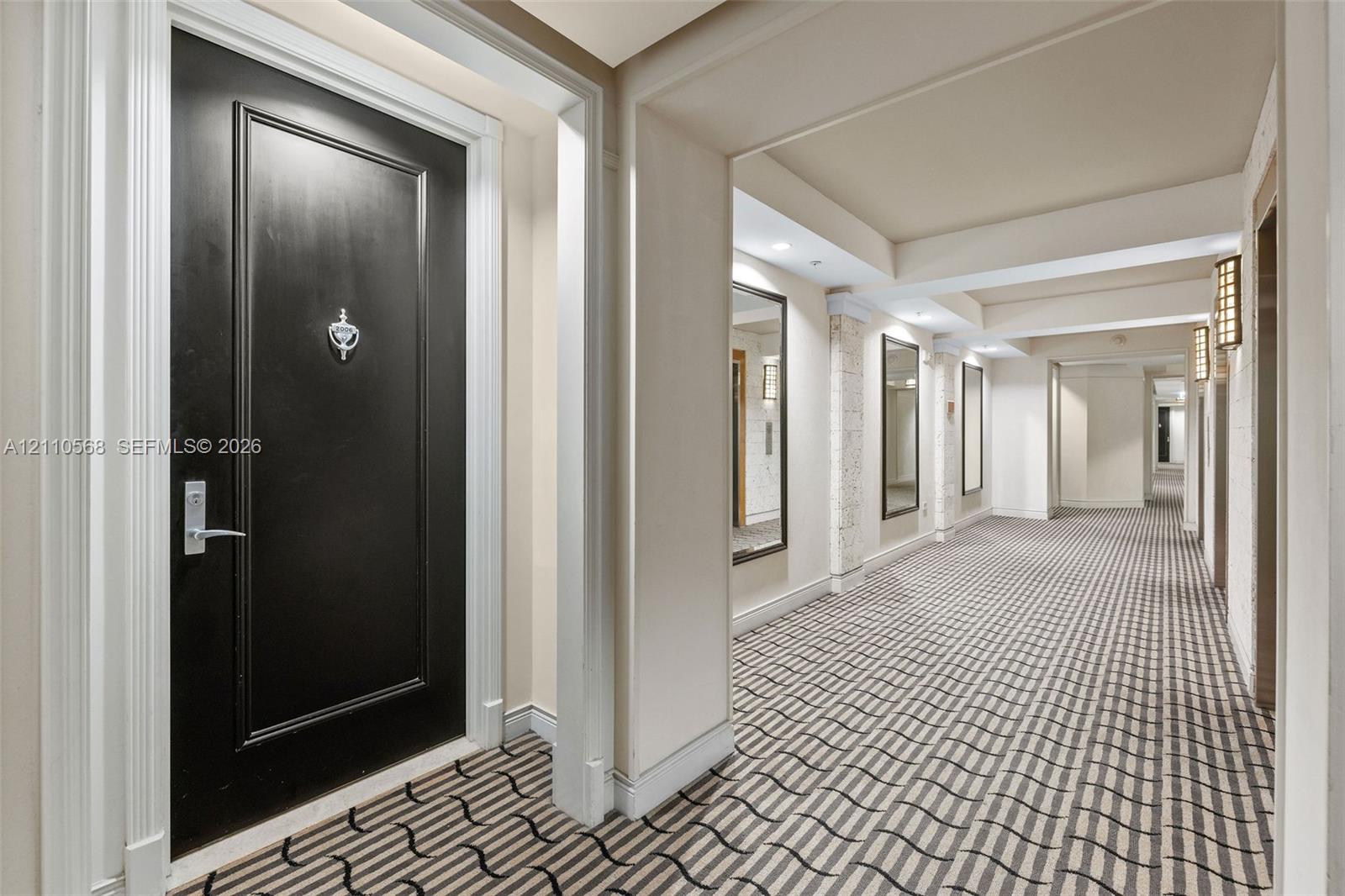 Black paneled entry door with chrome hardware, patterned wall-to-wall carpeting, recessed ceiling lighting, wall-mounted rectangular mirrors, and architectural pillars