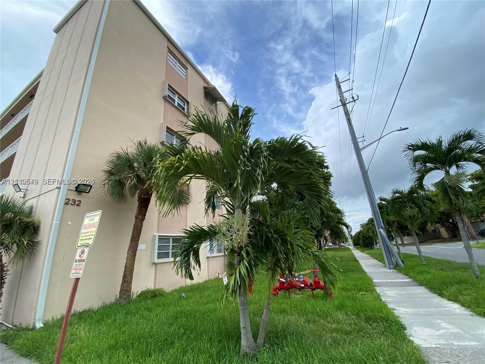 Multi-story beige exterior featuring horizontal windows, concrete sidewalk, palm landscaping, and grass lawn